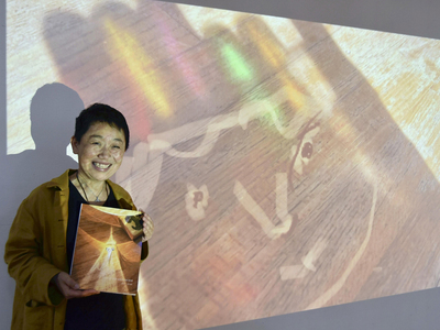作品を基にした絵本を手にする小原さん。背後の壁に投影された作品は、カラフルな持ち手のドライバーセットの影を写した写真に顔を書き込んだ